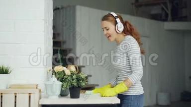 漂亮的女孩在家里擦桌子，通过耳机听音乐，带着快乐唱歌跳舞